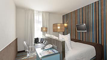 A hotel room with a bed, a desk with a telephone and a magazine, a chair, and a television mounted on the wall.