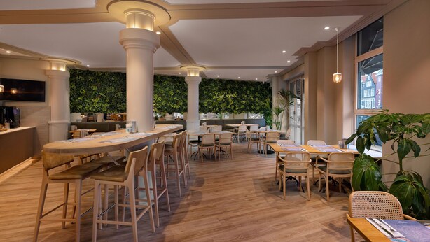 A modern restaurant with wooden tables and chairs, a green wall, and large windows. - WestCord City Centre Hotel Amsterdam (Amsterdam)