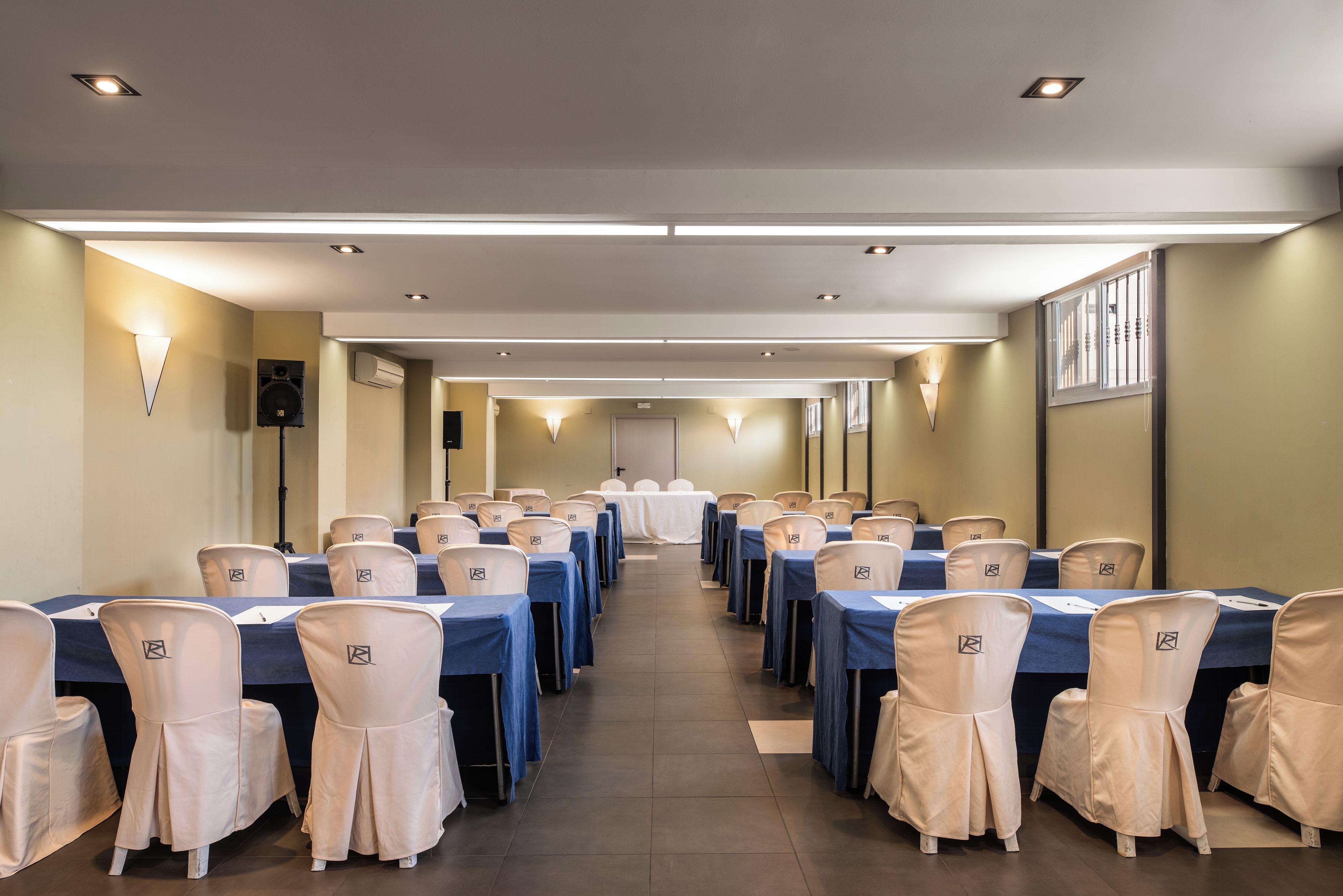 Un salón de conferencias con filas de mesas y sillas, equipado con luces de techo y altavoces montados en la pared.