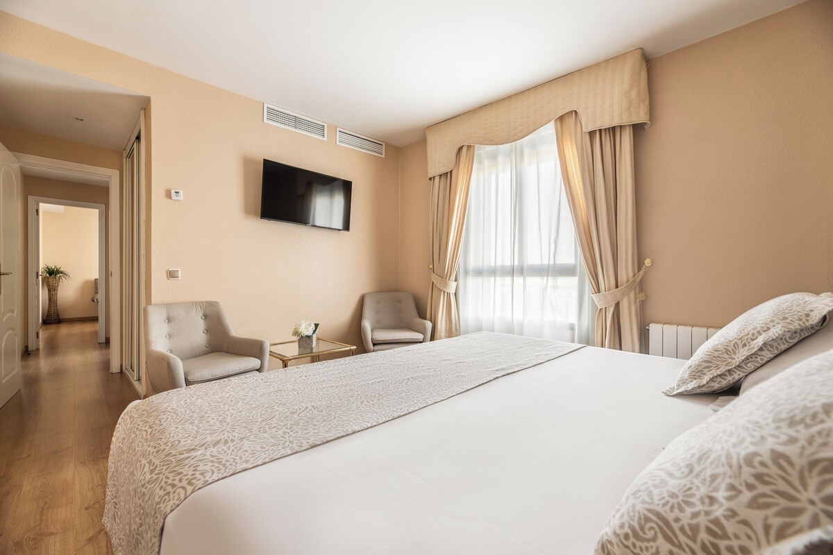 Hotellihuone, jossa on suuri sänky, kaksi nojatuolia, seinälle asennettu televisio ja ikkunasta avautuva näkymä ulos.