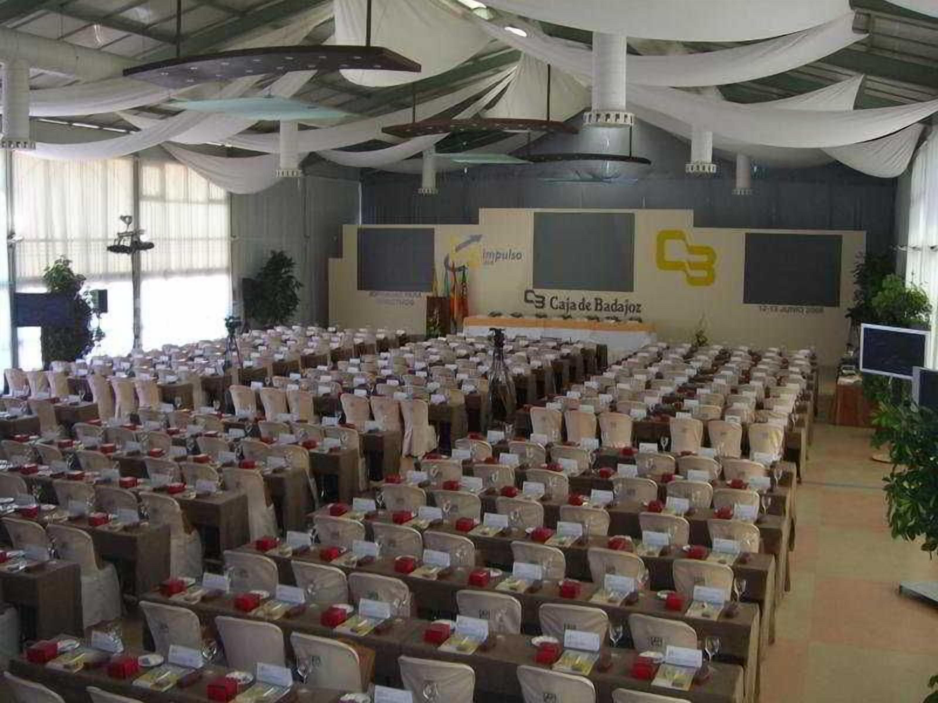 A conference room with rows of tables and chairs, a stage with a banner, and a large screen.