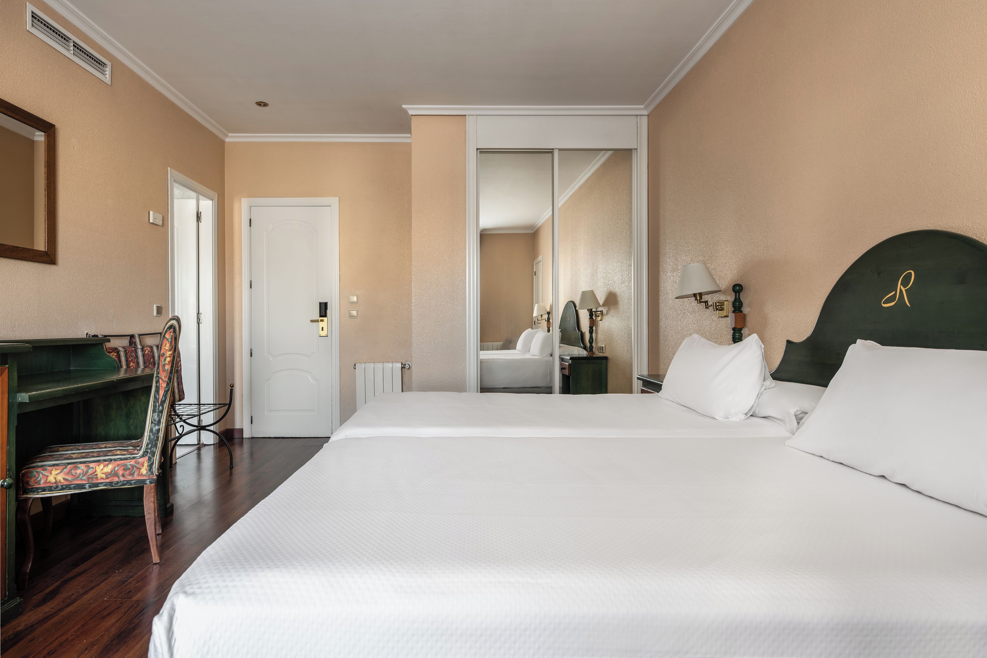 A hotel room with a large bed, a desk with a chair, a mirror, and a radiator.