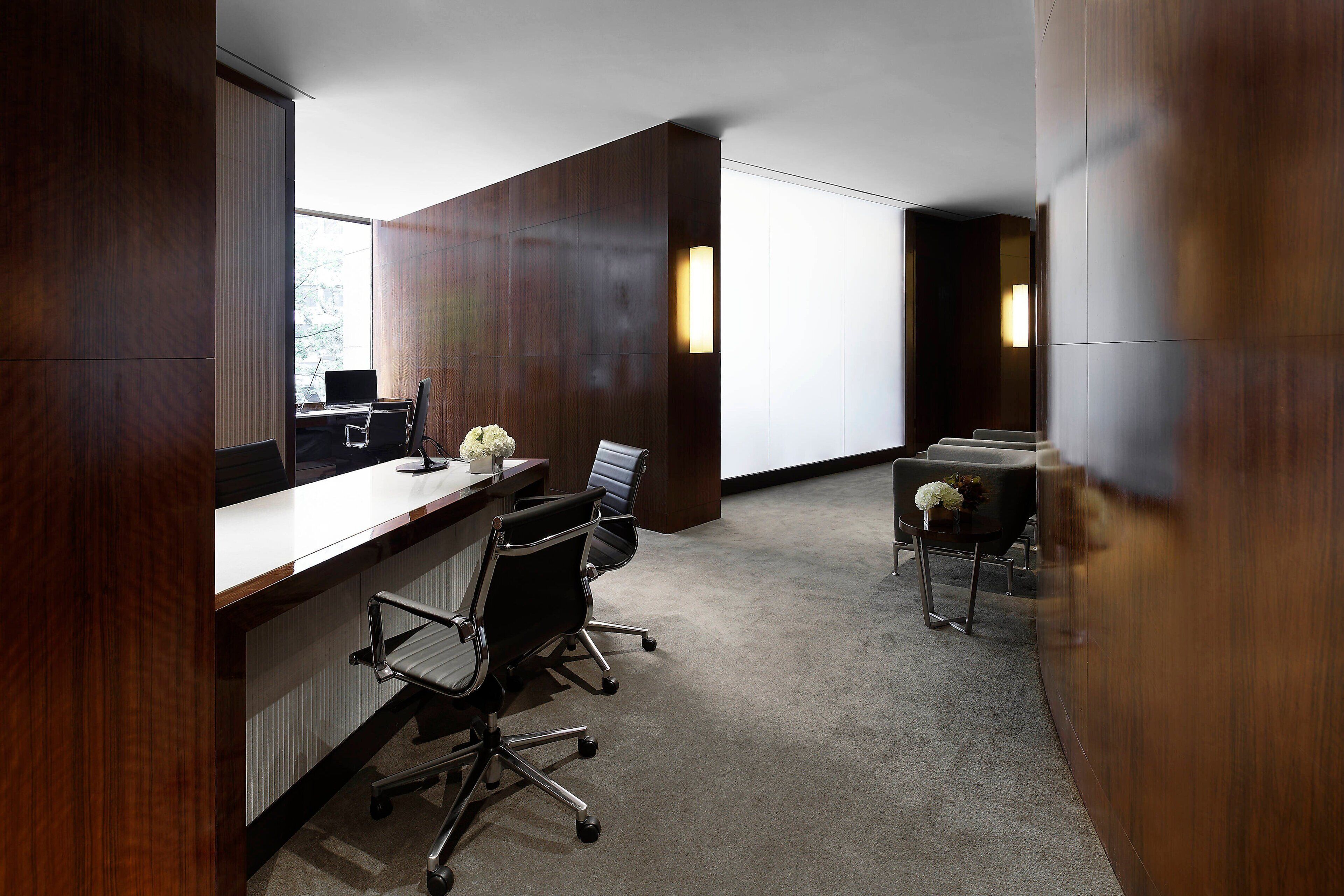A modern office space with a long wooden desk, multiple chairs, and a large window providing natural light.
