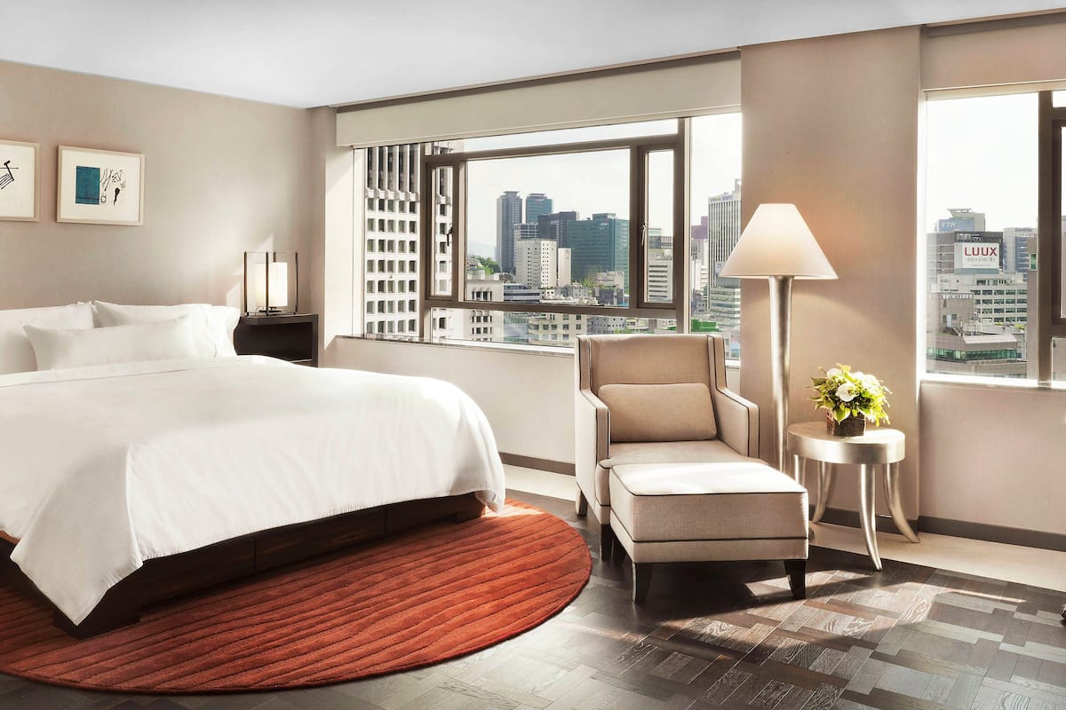 Hotelkamer met groot bed, stoel, klein tafeltje met lamp, en uitzicht op de stad door grote ramen.