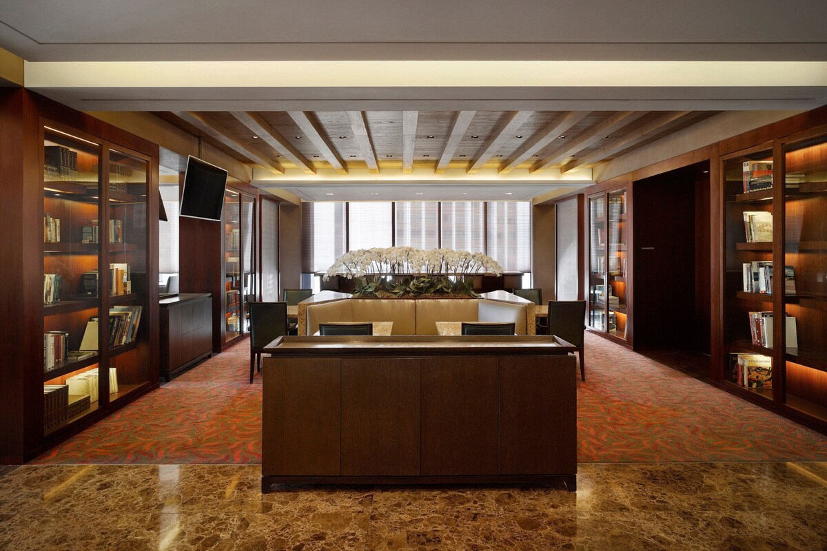 A hotel lobby with a central reception desk, bookshelves, and a large floral arrangement.