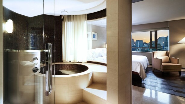 Een moderne hotelkamer met een glazen douchecabine, een groot bad en een bed, met uitzicht op de stad.