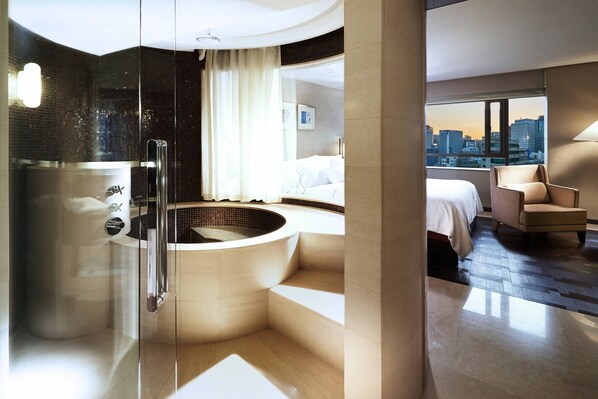 Een moderne hotelkamer met een glazen douchecabine, een groot bad en een bed, met uitzicht op de stad.