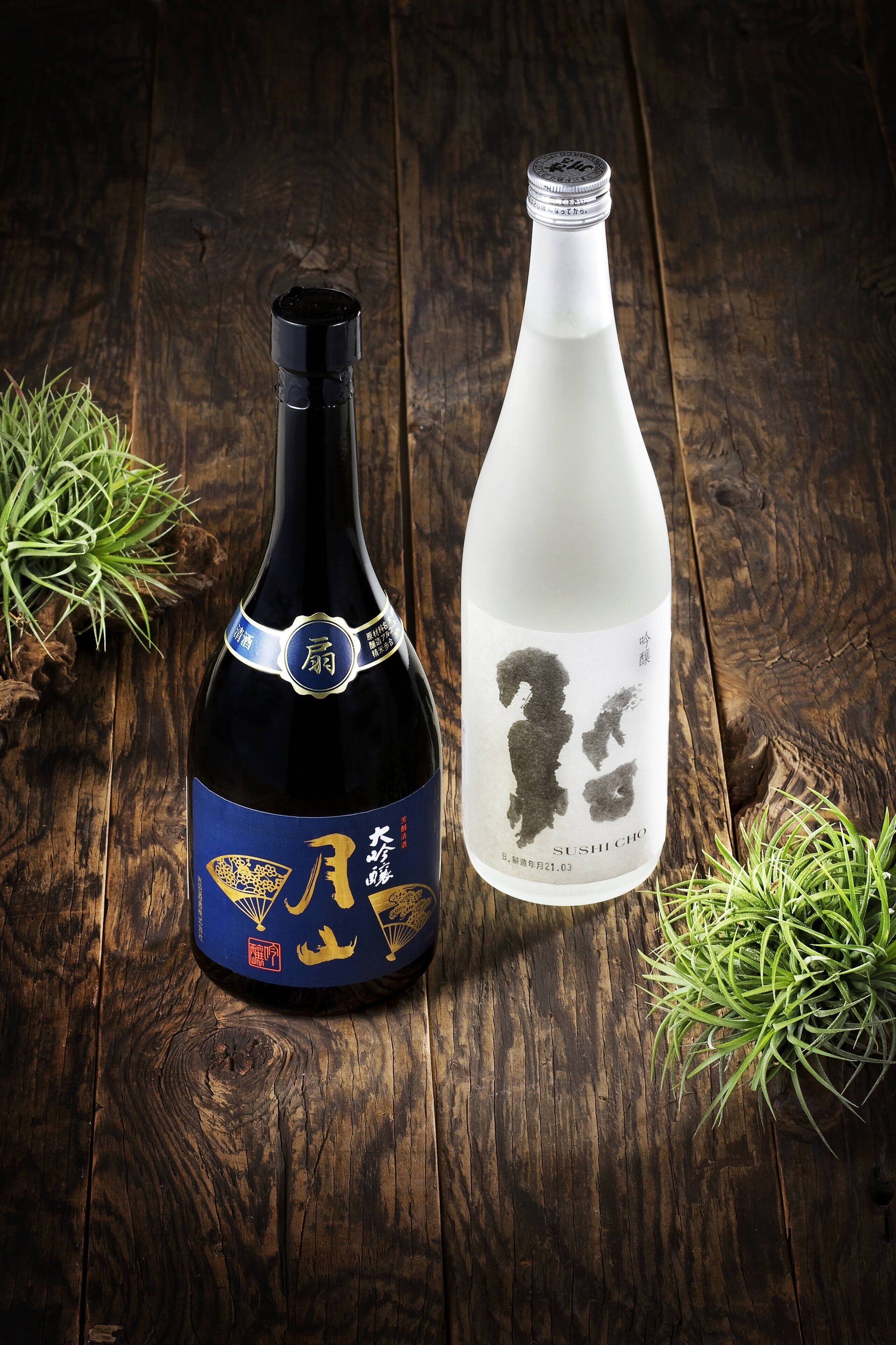 Two bottles of sake on a wooden surface with green plants.