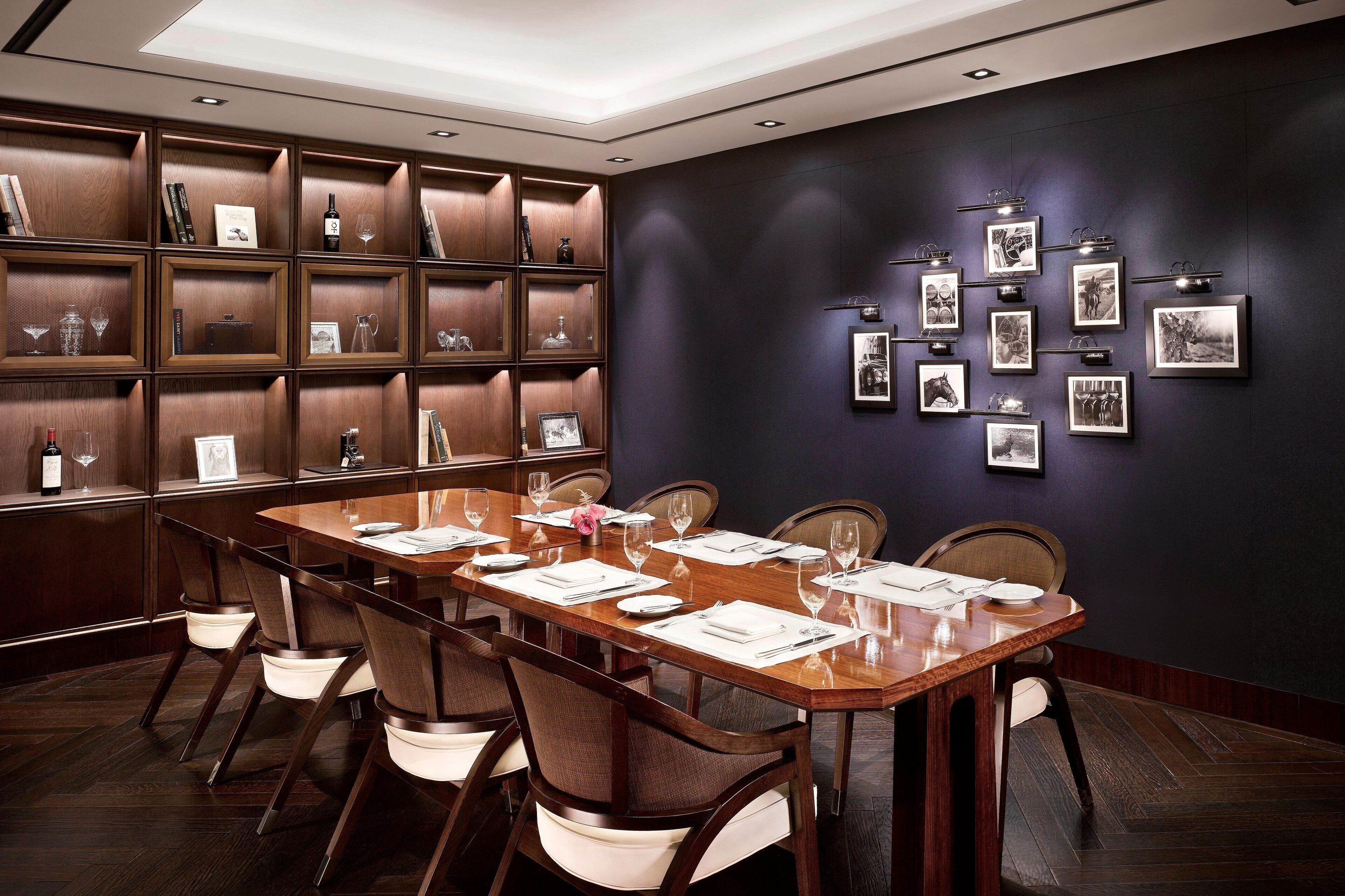 A conference room with a long table, chairs, a dark wall with framed pictures, and a wooden shelving unit with various items.