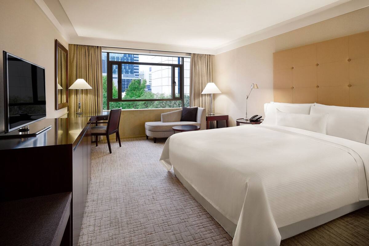 Een hotelkamer met een groot bed, een bureau met een televisie, een stoel, een klein tafeltje en een raam met uitzicht op gebouwen en bomen.