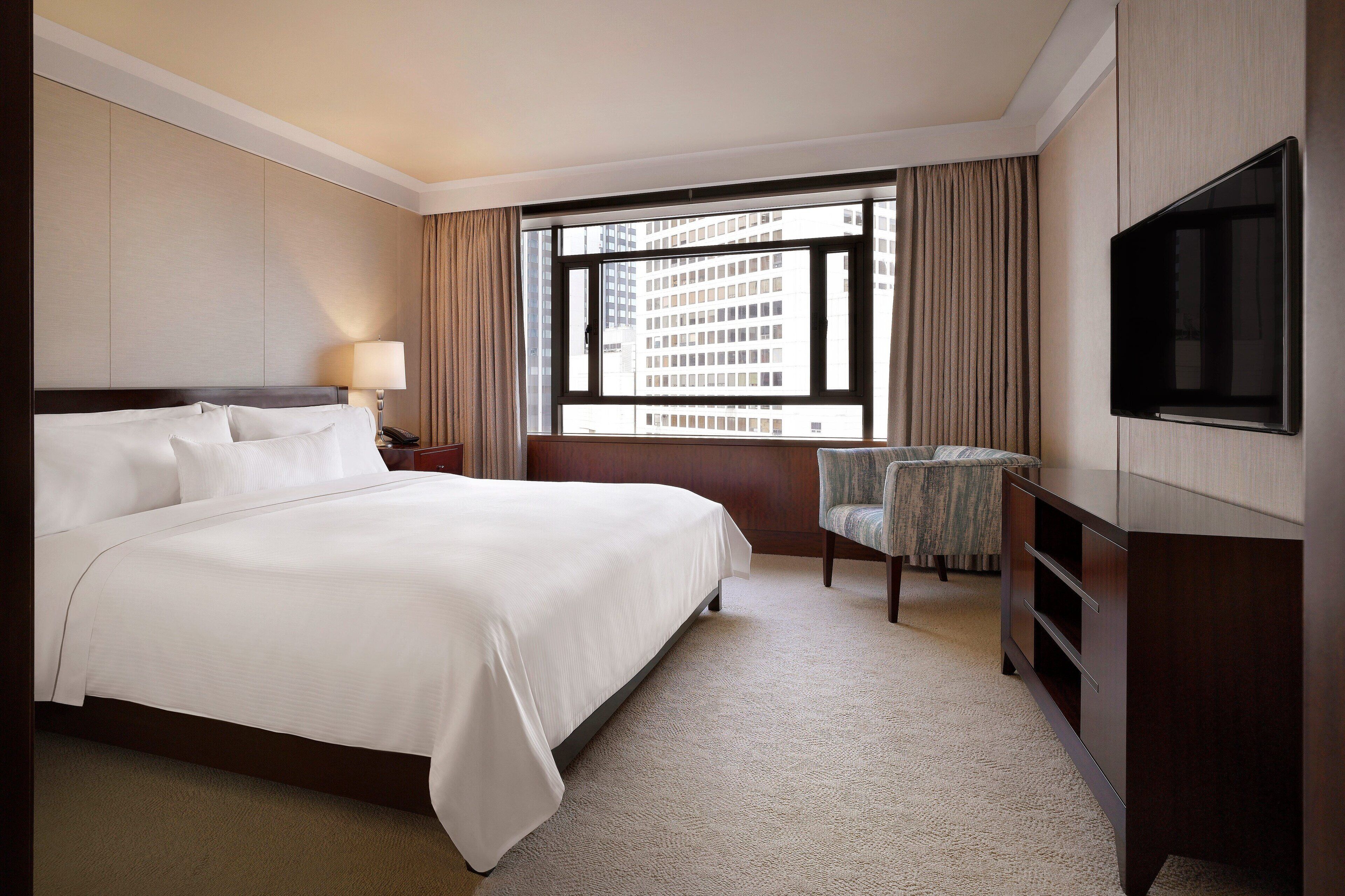 A hotel room with a large bed, a TV, a chair, and a window with a city view.