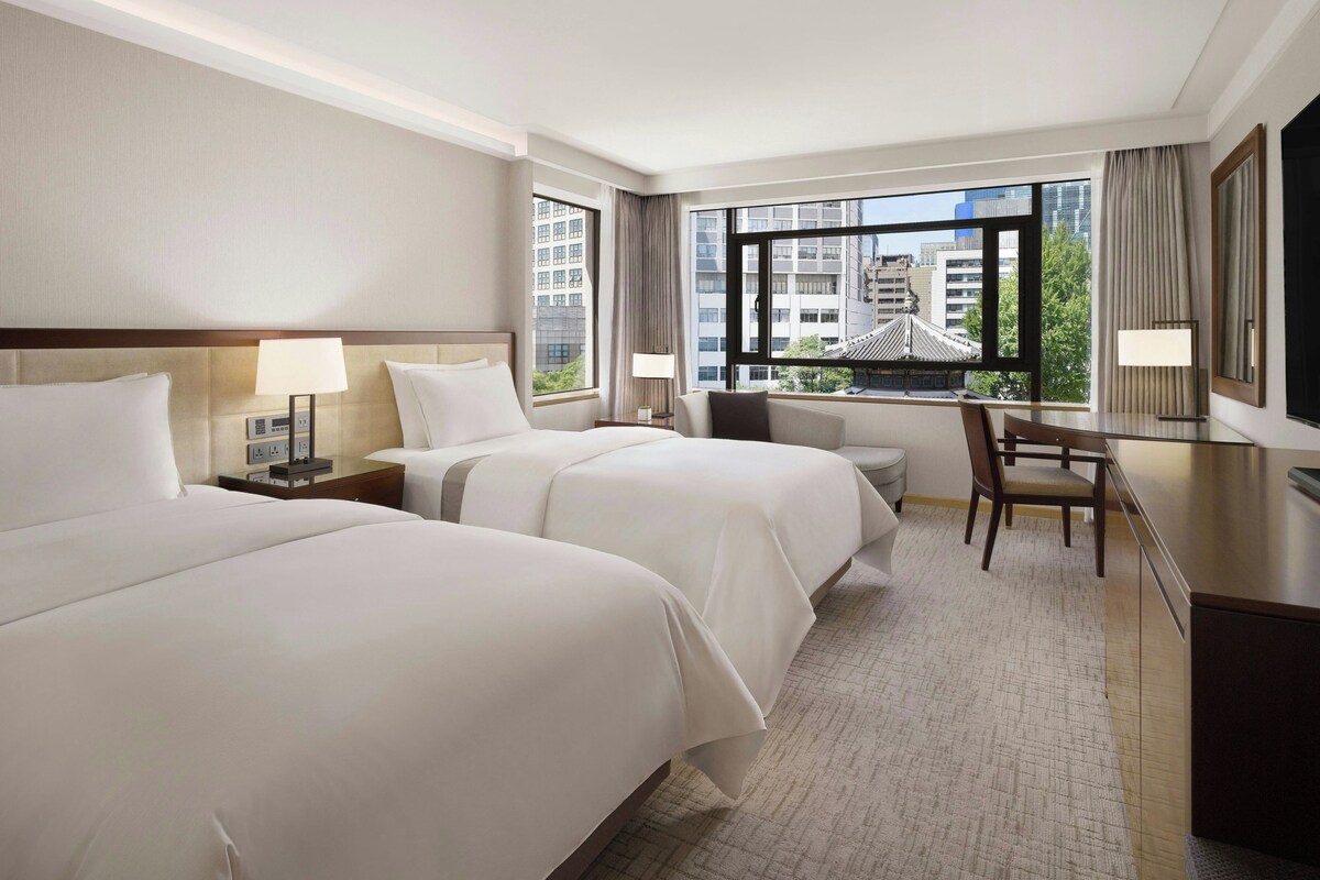 A hotel room with two beds, a desk, a chair, and a large window with a city view.