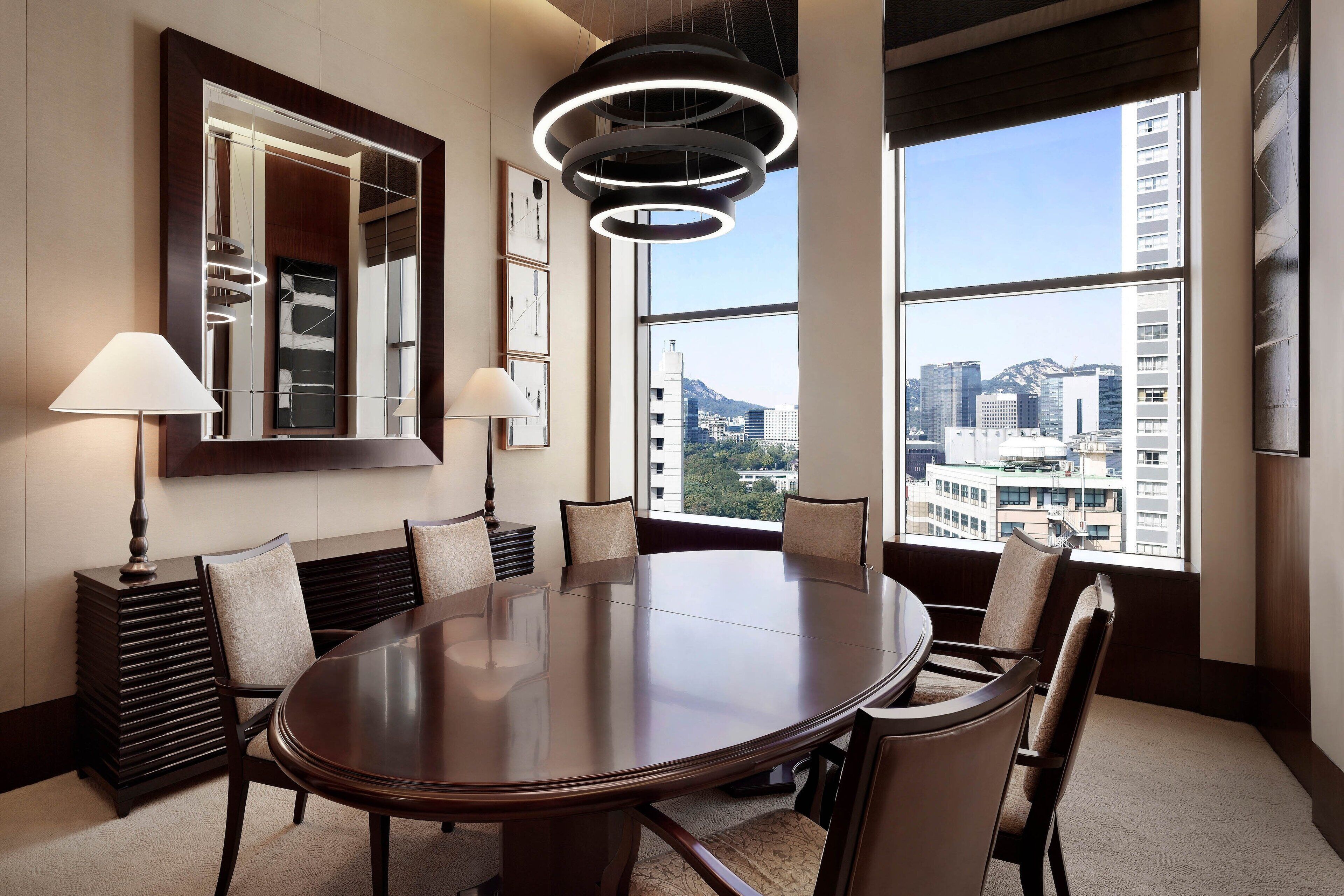 A conference room with a round table, chairs, a lamp, and a large window with a city view.