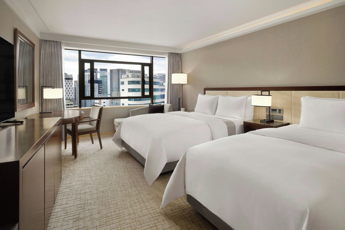 A hotel room with two beds, a desk, a chair, a TV, and a window with a city view.