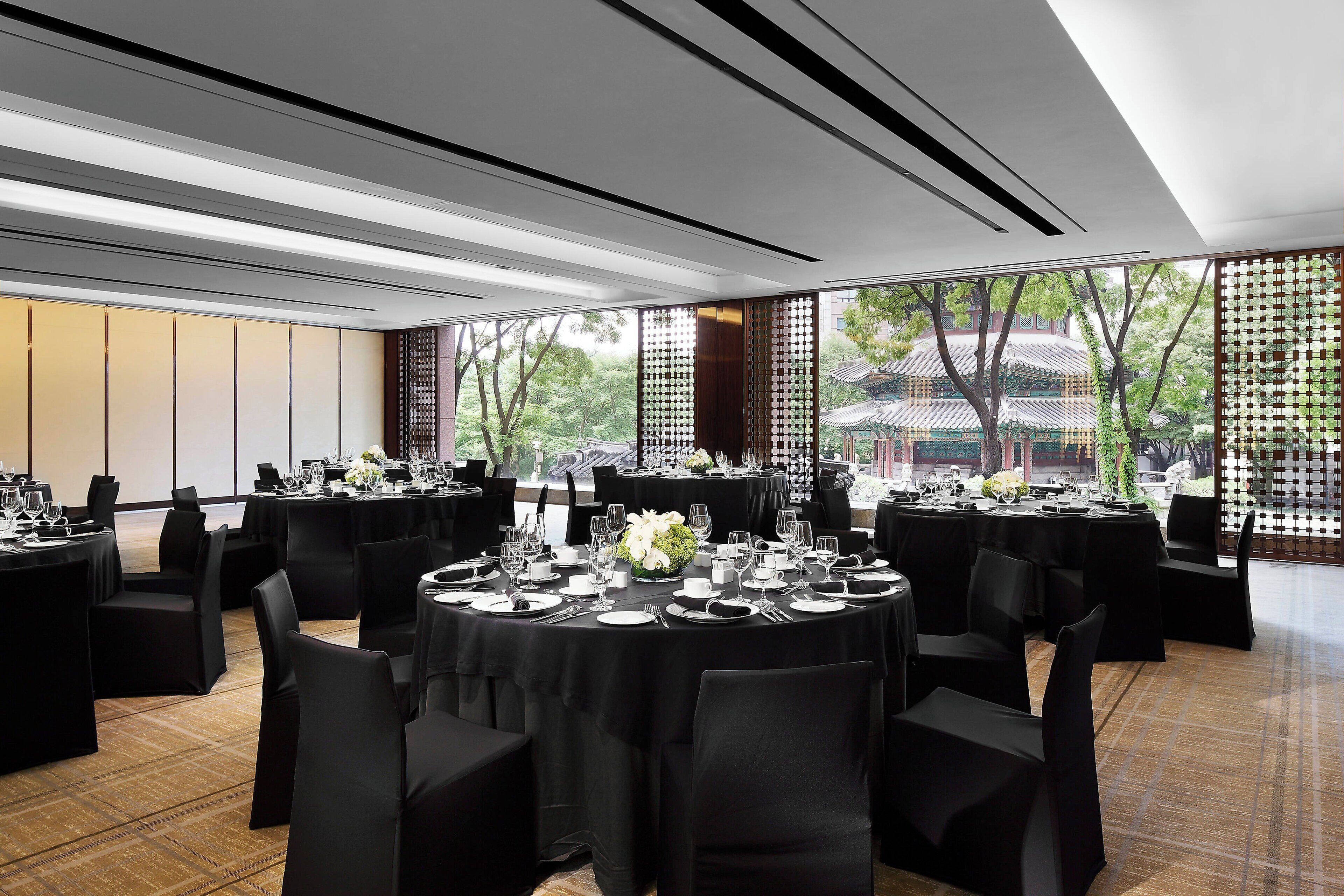 Een balzaal met ronde tafels gedekt voor een diner, grote ramen met uitzicht op een traditioneel gebouw, en een modern plafondontwerp.
