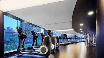 Uma academia com esteiras e equipamentos de exercício, grandes janelas com vista para a cidade, e teto curvo.