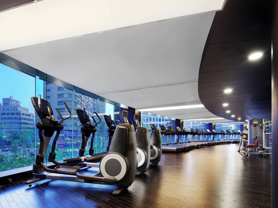 Uma academia com esteiras e equipamentos de exercício, grandes janelas com vista para a cidade, e teto curvo.