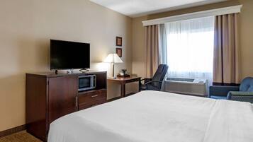 A hotel room with a bed, a desk with a TV, a lamp, a chair, and an air conditioning unit.