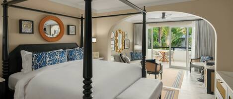 A four-poster bed with a canopy, a round mirror, and blue patterned pillows. There is a bench with a striped cushion, a dining area with a table and chairs, and a view of the outdoors through a glass door.