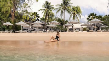 Una spiaggia con lettini e ombrelloni, una donna seduta sulla sabbia e palme sullo sfondo.