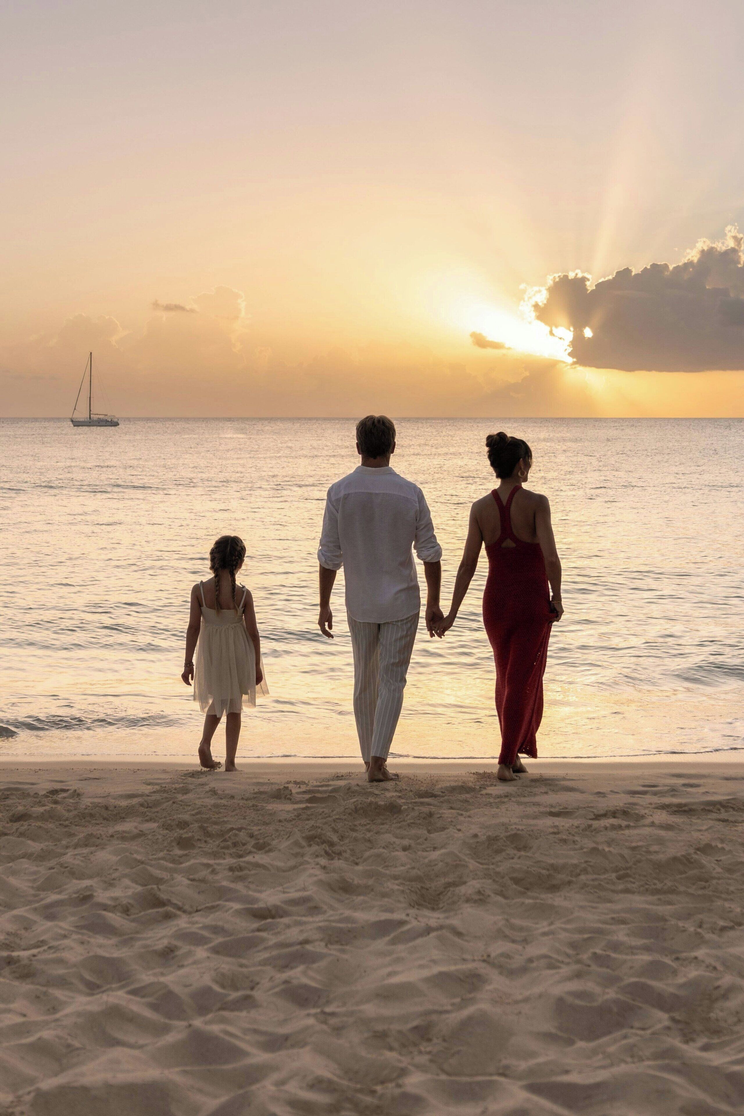 A family walking on a beach at sunset with a sailboat in the distance.