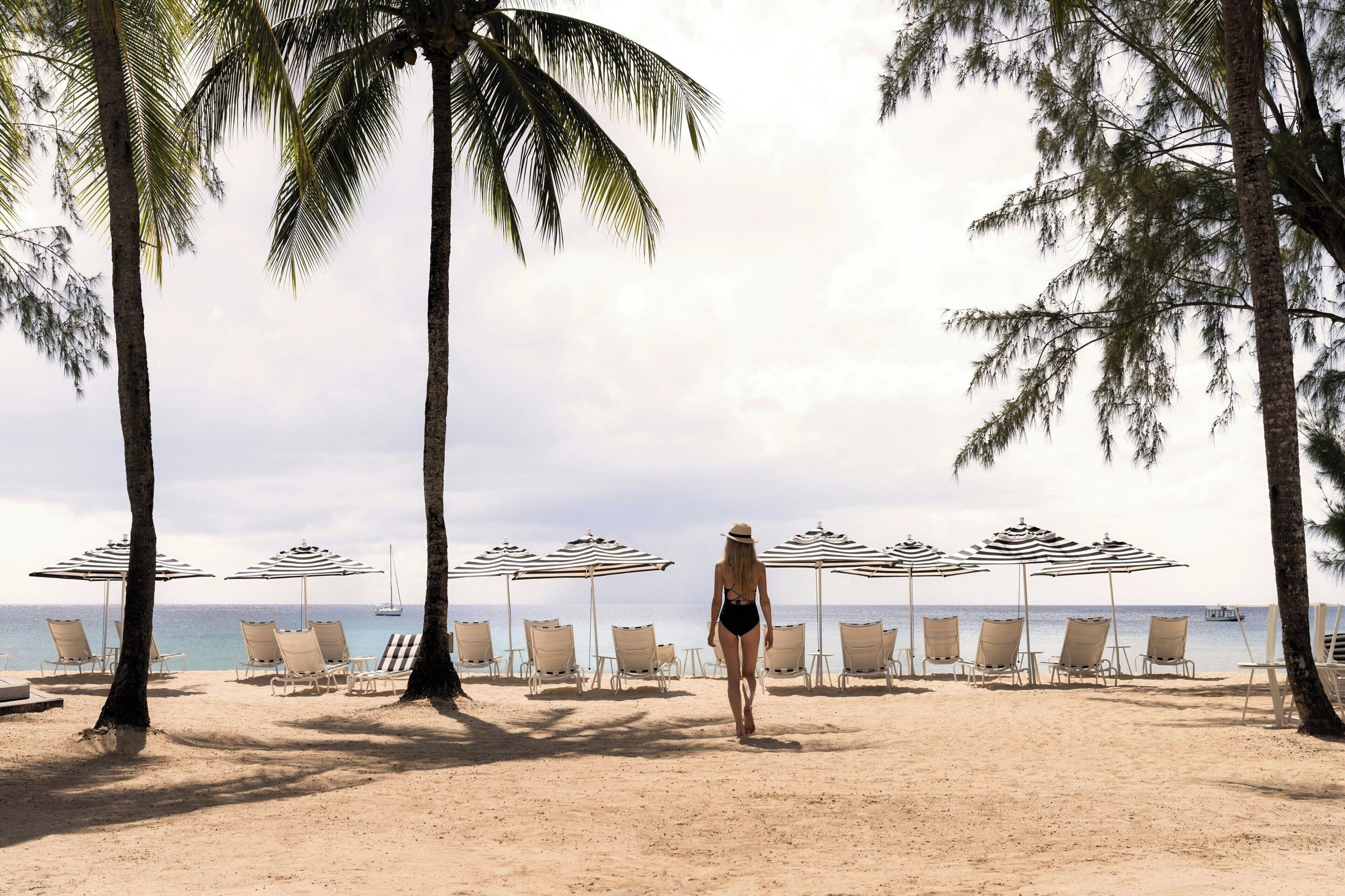 Una spiaggia con file di lettini e ombrelloni a righe, una persona che cammina e palme.