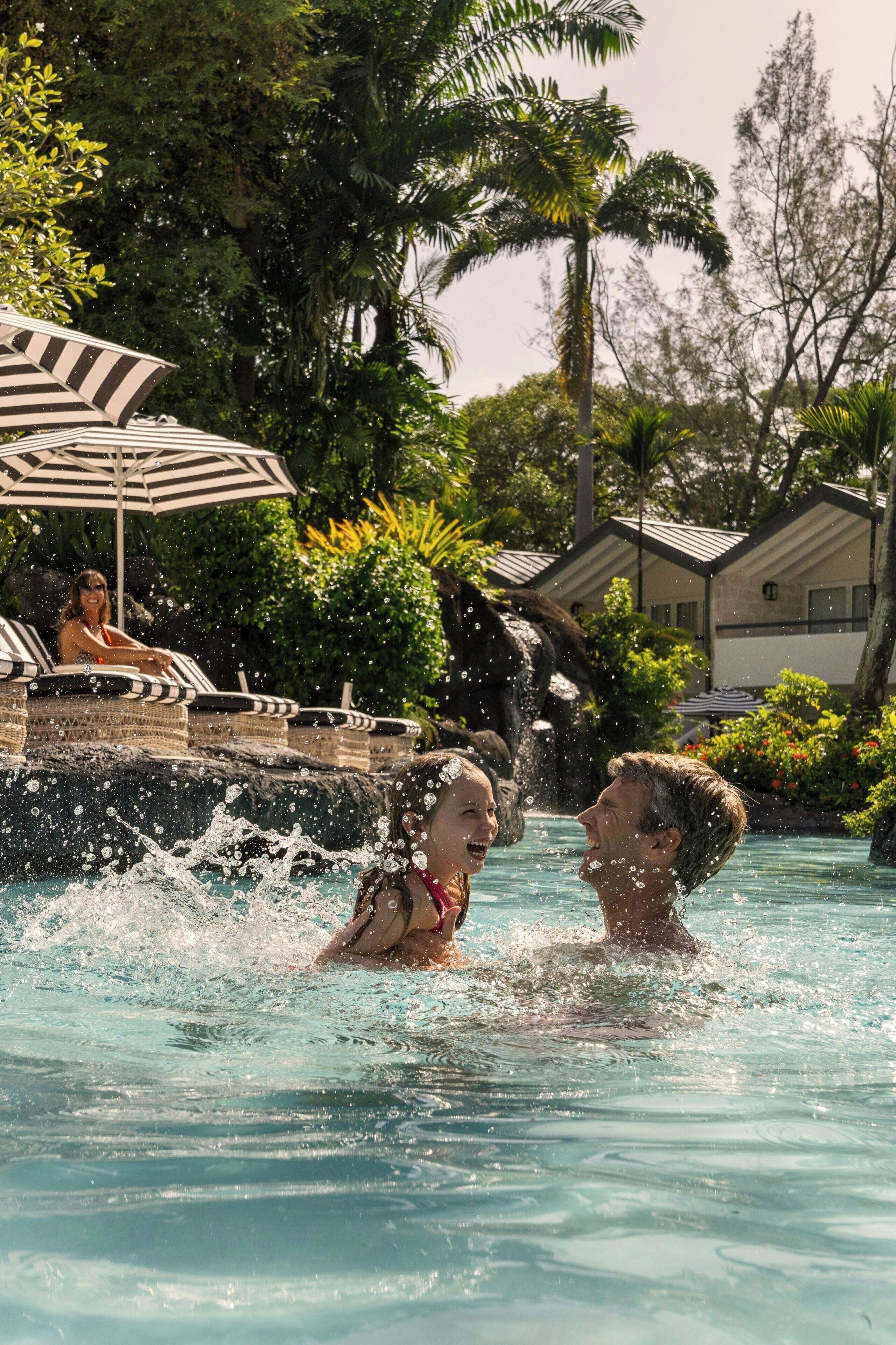 A pool area with lounge chairs and umbrellas, surrounded by lush greenery and a waterfall feature.