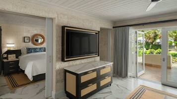 Habitación de hotel con una amplia televisión de pantalla plana en la pared, suelo de mármol, cama con cubrecama blanca, mesita de noche negra con lámpara, mueble de madera con cajones y puerta corredera de cristal que da a un balcón con vegetación.