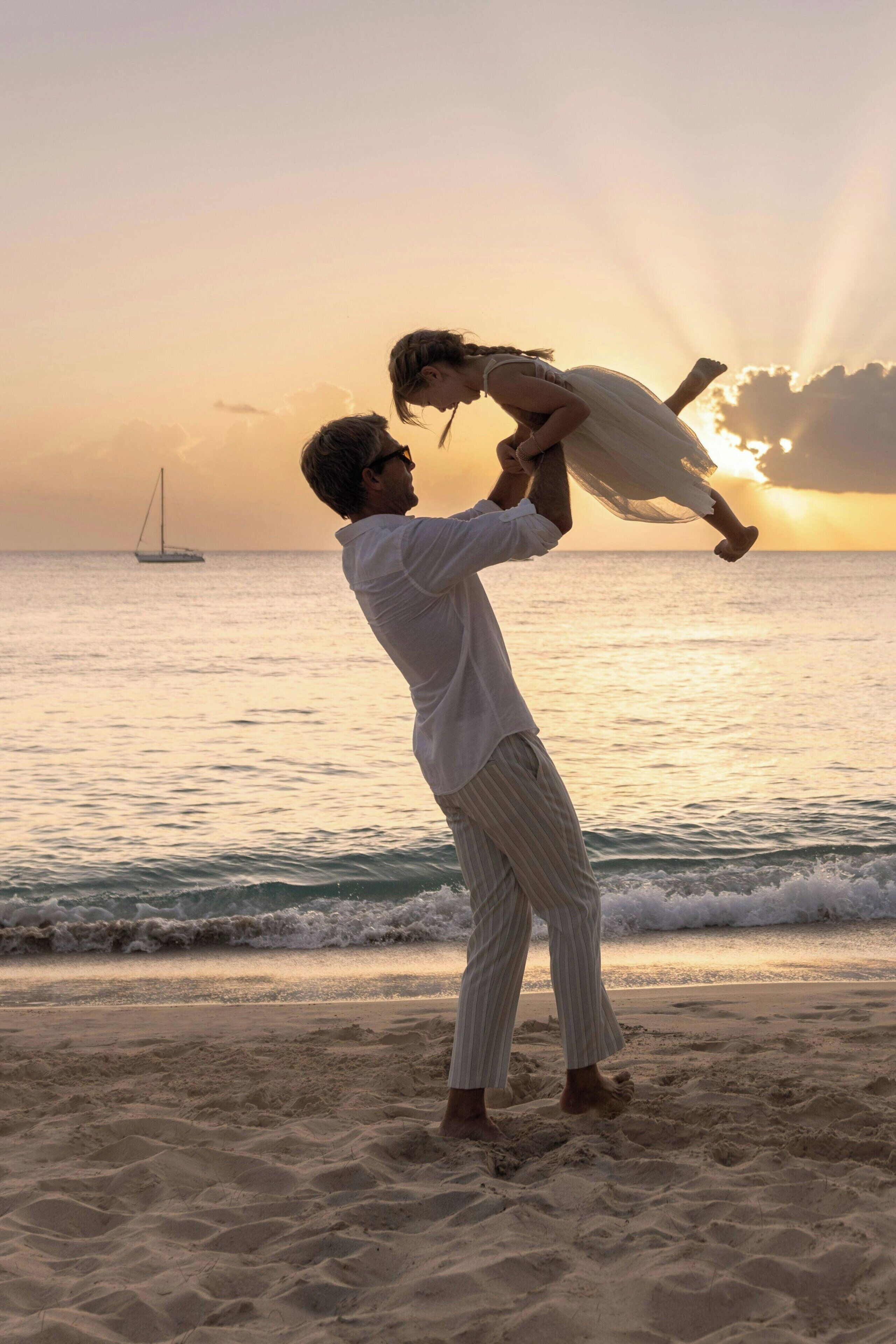 A person holding a child on a beach with a sailboat in the background during sunset.