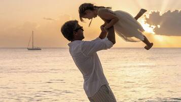 A person holding a child on a beach with a sailboat in the background during sunset.