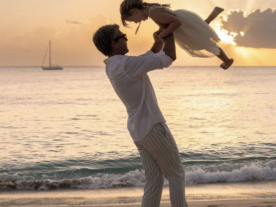 A person holding a child on a beach with a sailboat in the background during sunset.