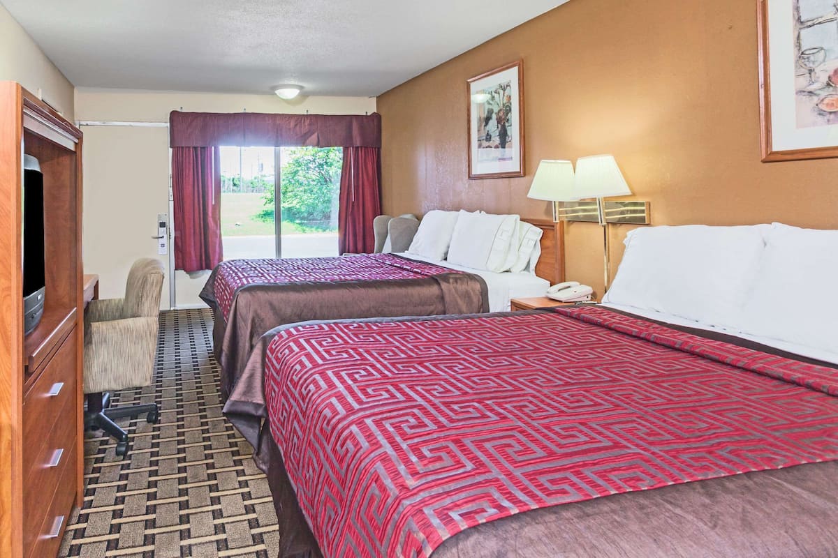 A hotel room with two beds, a desk, a chair, a lamp, and a view of the outdoors.