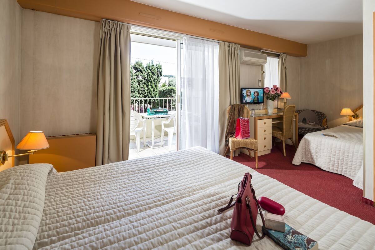 A hotel room with two beds, a desk with a TV, a balcony with a table and chairs, and a view of trees.