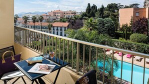 A balcony with a view of a swimming pool, palm trees, and buildings in the background. - Beausejour (Cannes)