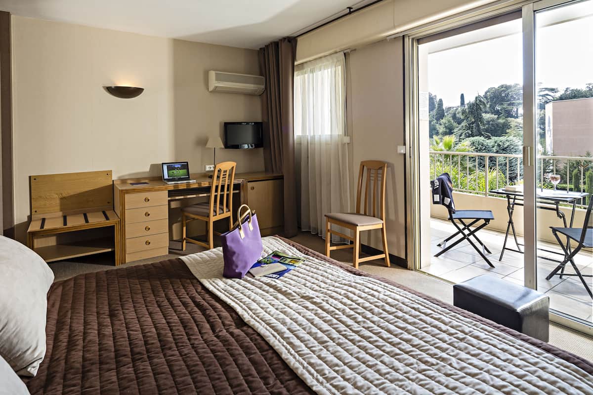 A hotel room with a bed, a desk with a laptop, a TV, and a balcony with a table and chairs.