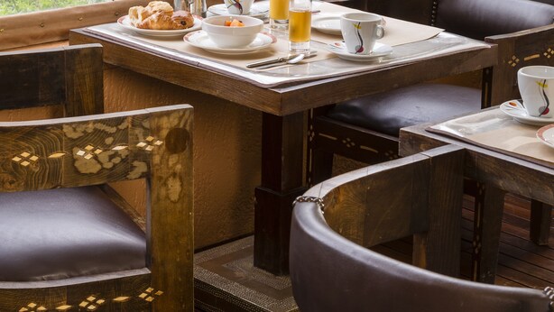 A dining area with wooden tables and chairs, a view of greenery outside, and a breakfast setting with plates, cups, and a glass of orange juice. - Beausejour (Cannes)