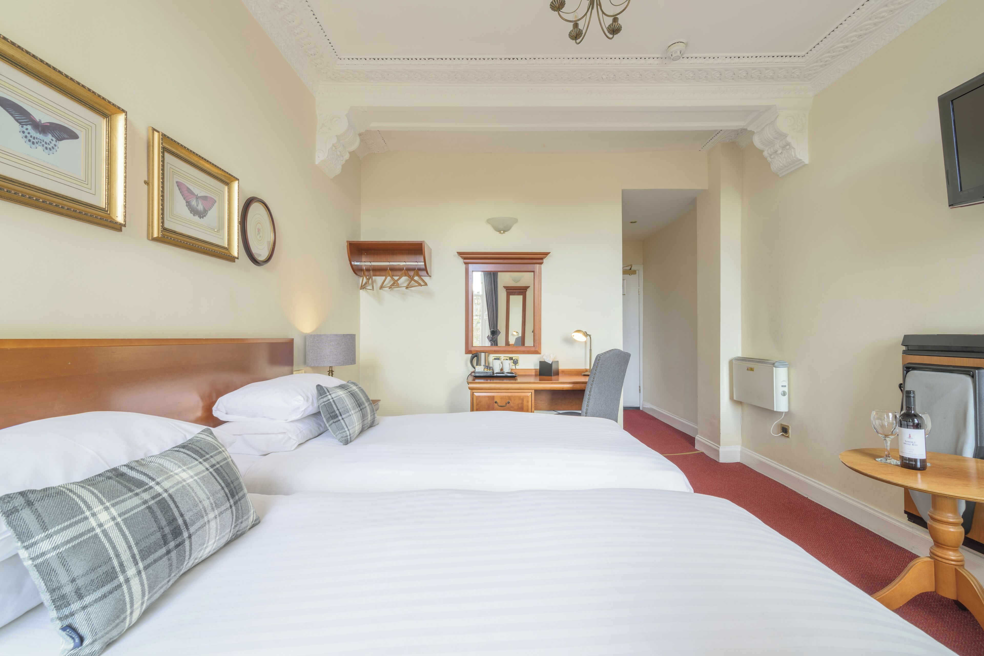 A hotel room with two beds, a desk, a chair, a mirror, and a small table with a bottle and glass.