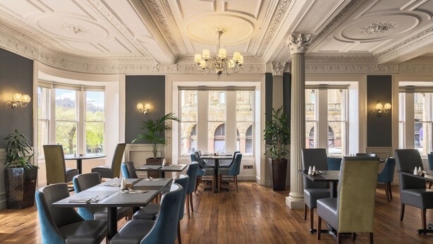 A dining area with wooden flooring, tables, and chairs, large windows with a view, and decorative ceiling details. - Old Waverley Hotel (Edinburgh)