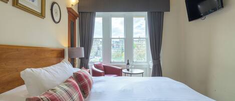 A hotel room with a large bed, a television mounted on the wall, a small table with a bottle, and a view of the city through the window.