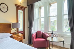A hotel room with a bed, a nightstand with a lamp, a red armchair, a small table with wine glasses, and a view of the city through large windows.