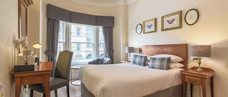 A hotel room with a large bed, a desk with a telephone, a chair, a window with curtains, and a chandelier.