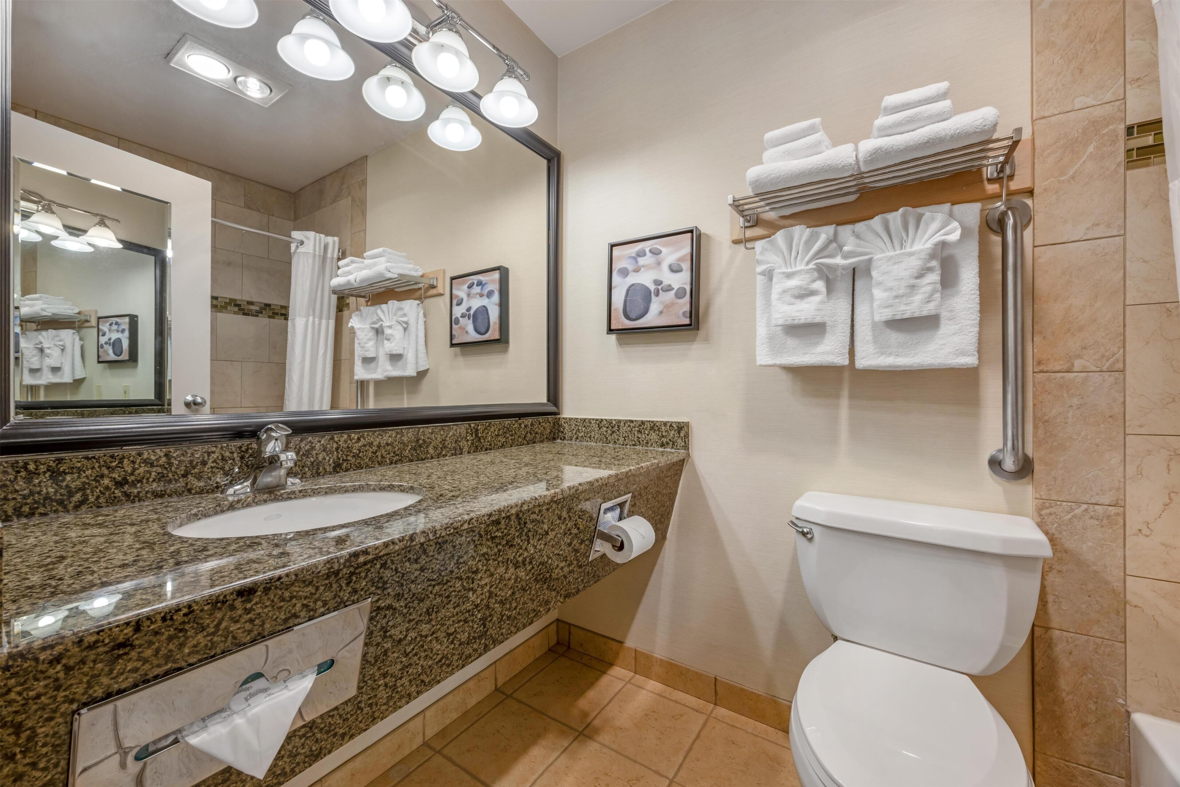 A bathroom with a granite countertop sink, a white toilet, a towel rack with towels, a mirror, and a framed picture on the wall.