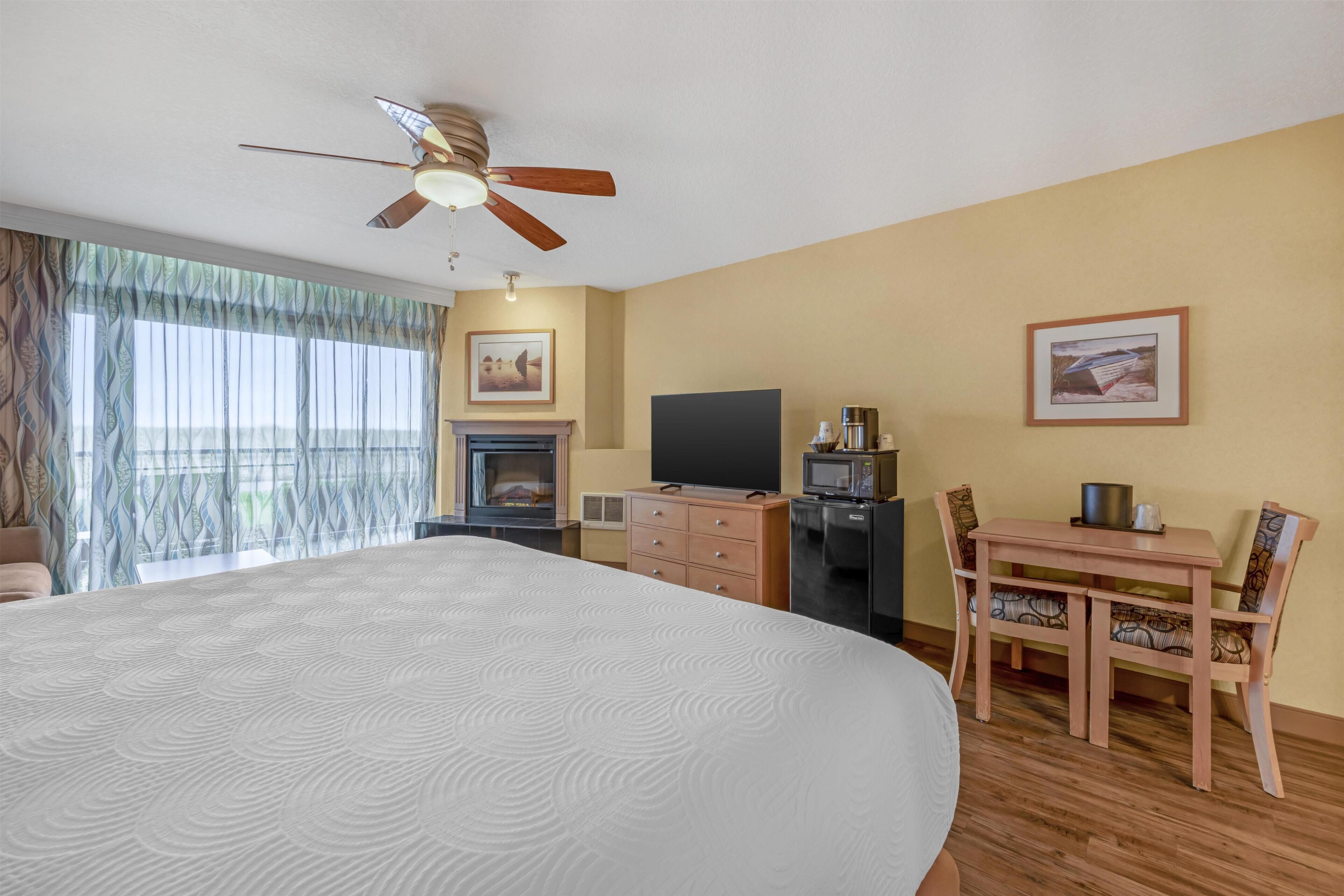Habitación de hotel con una cama grande, ventilador de techo, chimenea, televisión, escritorio con silla y ventana con cortinas.