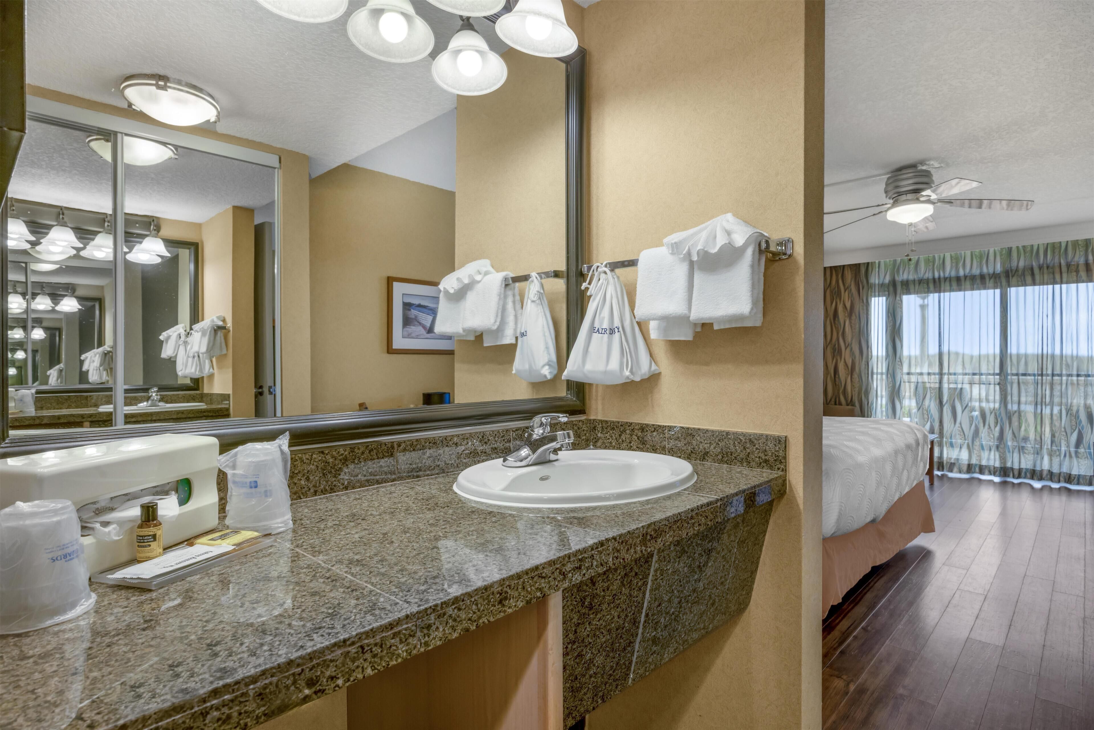 A hotel bathroom with a granite countertop sink, a large mirror, and a ceiling fan.