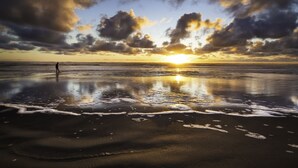 Uma praia com um pôr do sol, ondas calmas e uma pessoa parada na areia.