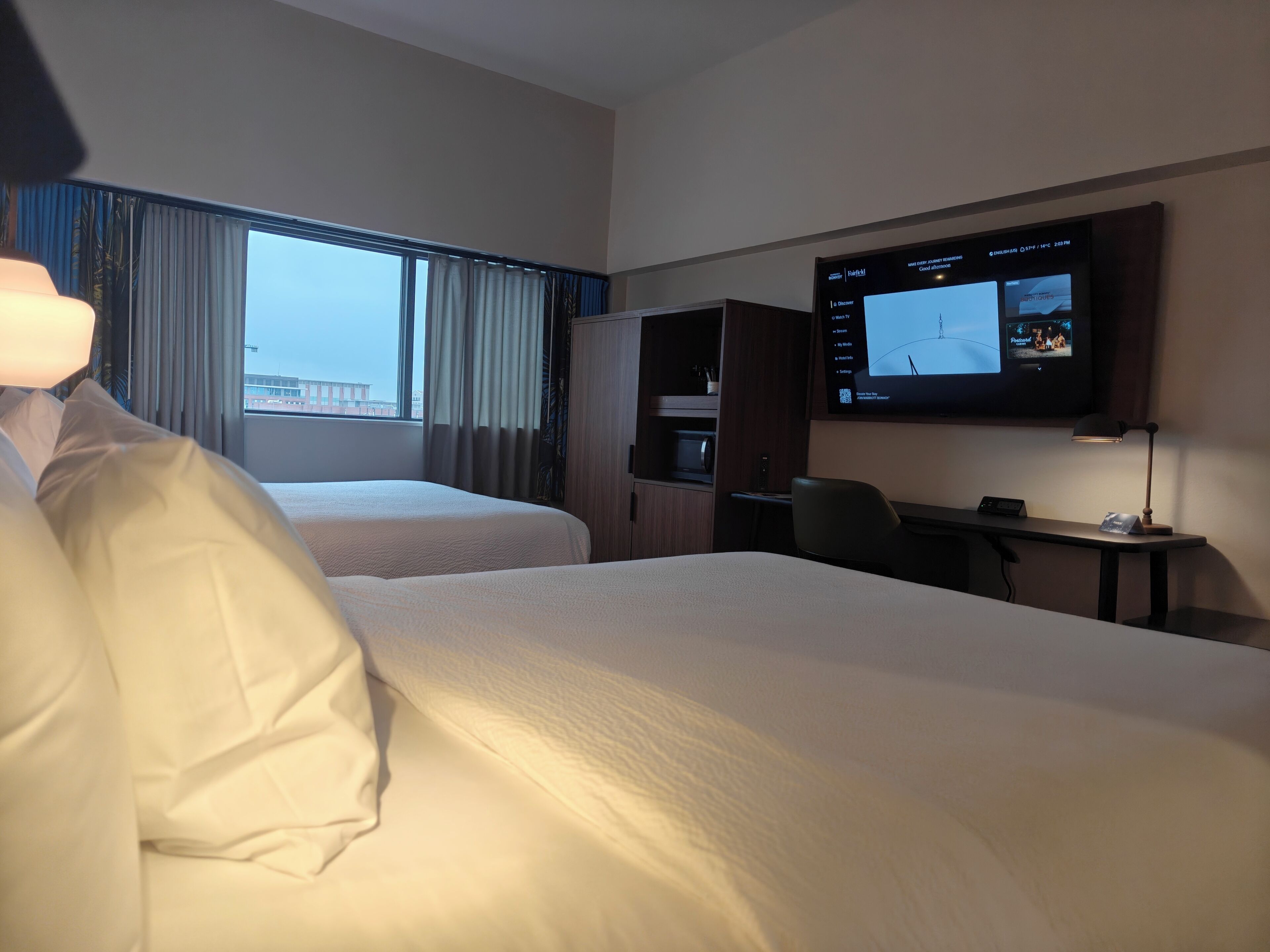 A hotel room with two beds, a desk, a TV, and a window with curtains.