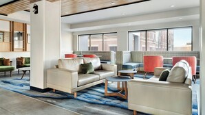 A modern hotel lobby with a beige sofa, armchairs, and a coffee table. There is a large window providing a view of the city.