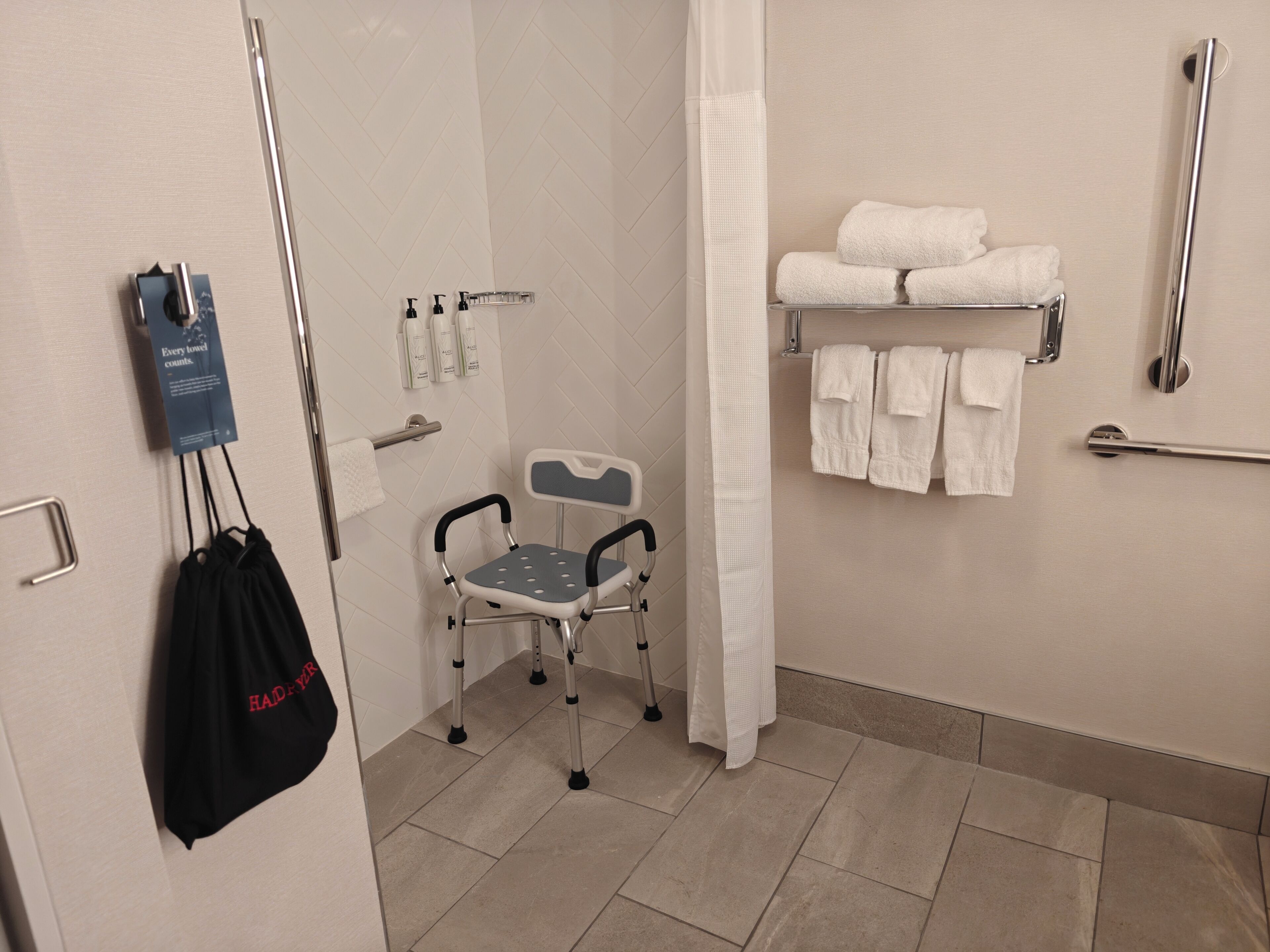 A bathroom with a shower, a grab bar, a wheelchair, a towel rack, and a bag hanging on the wall.