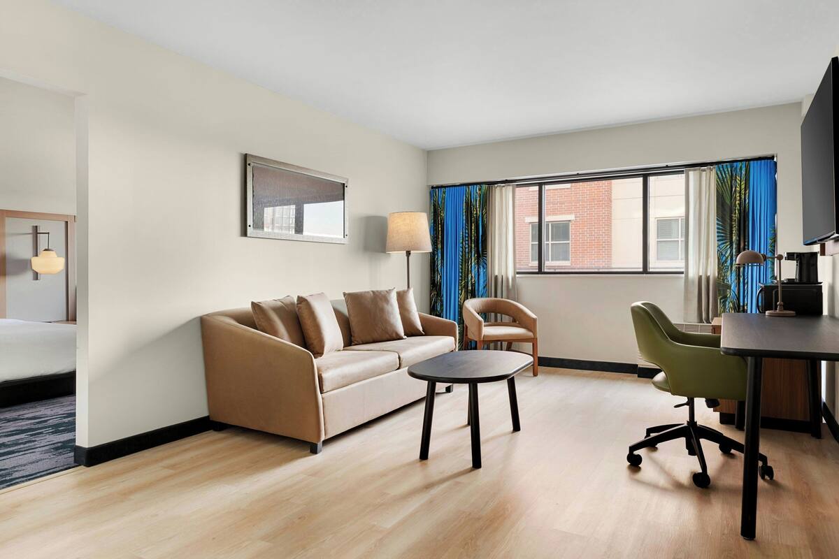 Ein Hotelzimmer mit einer Couch, einem kleinen Tisch, einem Stuhl, einem Schreibtisch und einem Fernseher.
