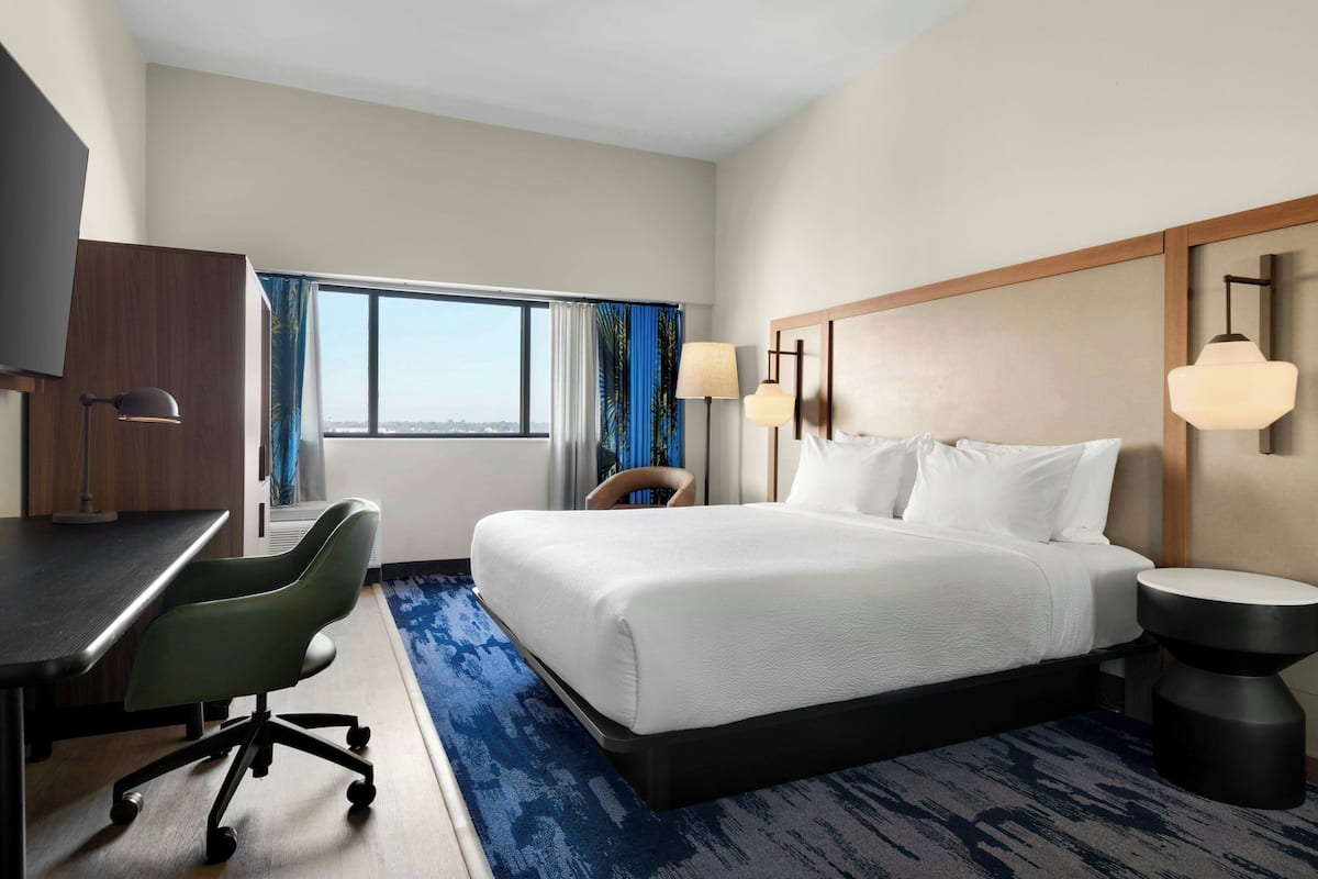 Ein Hotelzimmer mit einem großen Bett, einem Schreibtisch, einem Stuhl, einer Lampe und Blick auf die Stadt.